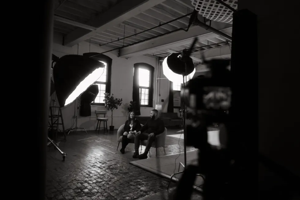A man and woman sit together on a couch in a well-lit studio, with cameras and lighting equipment visible in the foreground