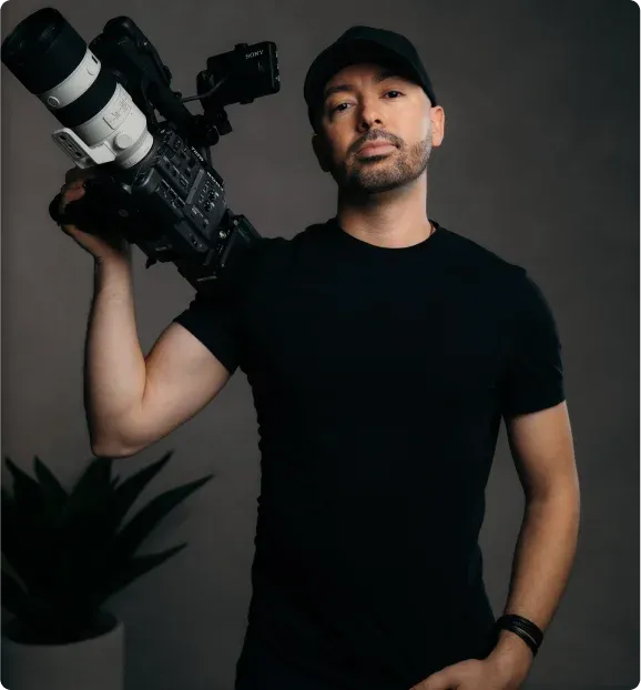 A person wearing a black t-shirt holds a professional camera rig, with a plant in the background against a gray backdrop