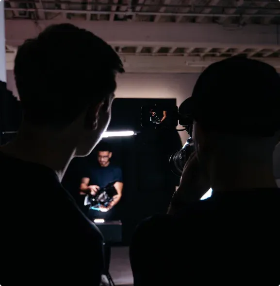 Two individuals observe a photoshoot, one holding a camera while the other is behind, with bright lighting and a dark backdrop