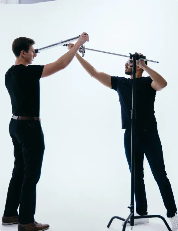 Two men in black shirts collaborate to adjust equipment on a stand in a bright, minimalist studio setting